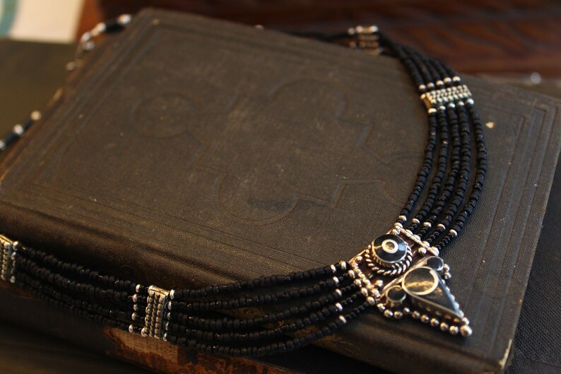 May include: A multi-strand necklace with black beads and silver-toned accents. The pendant features a geometric design with black and silver elements. The necklace is displayed on a dark, textured surface.