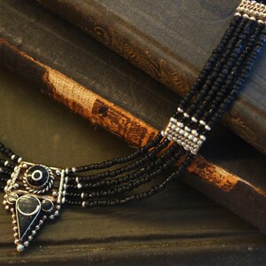 May include: A multi-strand black beaded necklace with a silver-toned pendant. The pendant has a geometric design with black accents. The necklace is displayed on a dark, textured surface, possibly a book.