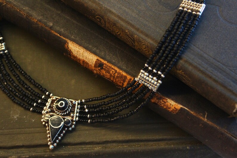 May include: A multi-strand black beaded necklace with a silver-toned pendant. The pendant has a geometric design with black accents. The necklace is displayed on a dark, textured surface, possibly a book.