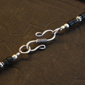 May include: Close-up of a necklace with a silver clasp and black beads. The necklace features a series of small, black, rectangular beads interspersed with silver-colored, round beads. The clasp is a silver-colored S-hook design.
