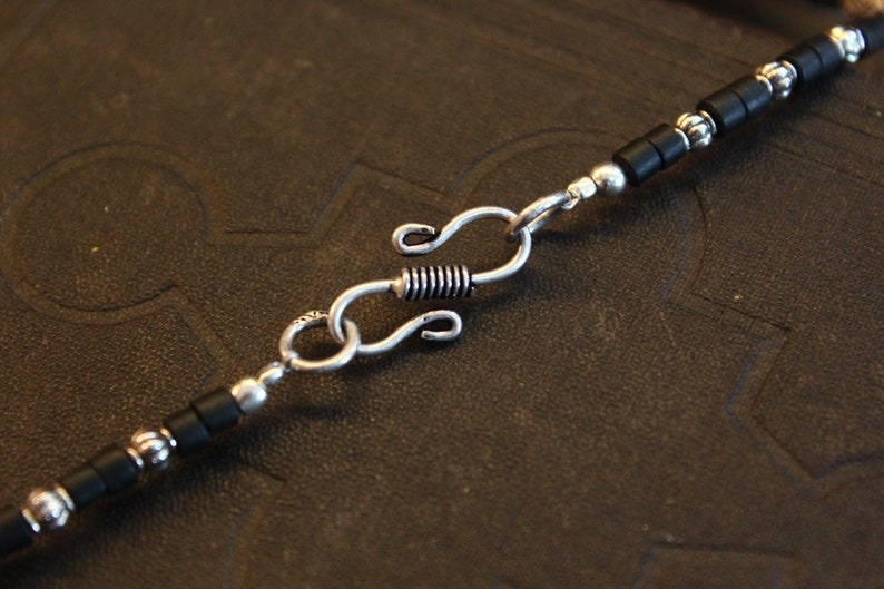May include: Close-up of a necklace with a silver clasp and black beads. The necklace features a series of small, black, rectangular beads interspersed with silver-colored, round beads. The clasp is a silver-colored S-hook design.