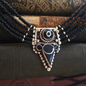 May include: A multi-strand black beaded necklace with a silver-toned pendant. The pendant features black and silver geometric shapes and a decorative border. The necklace is displayed against a backdrop of antique books.