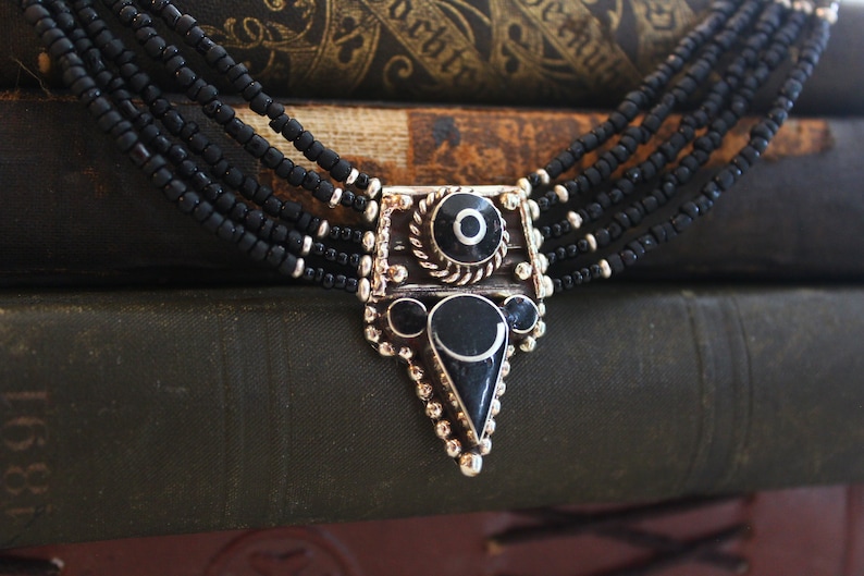 May include: A multi-strand black beaded necklace with a silver-toned pendant. The pendant features black and silver geometric shapes and a decorative border. The necklace is displayed against a backdrop of antique books.
