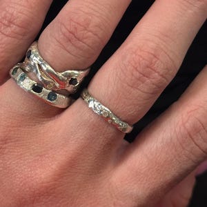 May include: Three silver rings are displayed on fingers. One ring has a textured, organic design with embedded blue and black gemstones. Another is a thin band with small, clear stones. The rings are on a hand.