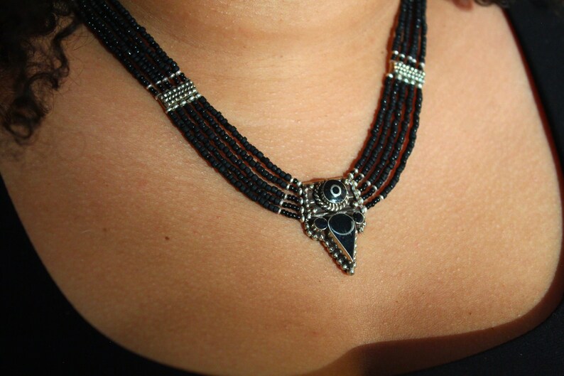 May include: A multi-strand black beaded necklace with a silver-toned pendant. The pendant has a central black stone and is surrounded by intricate silver detailing. The necklace is worn around the neck.
