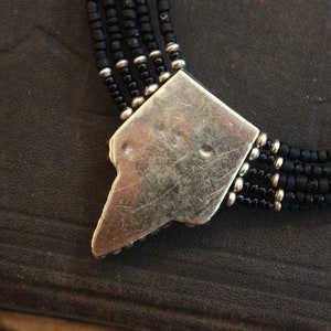 May include: Close-up of a black beaded necklace with a silver-colored clasp. The necklace features multiple strands of small, dark beads. The clasp is a geometric shape with a worn, textured appearance, and is connected to the beaded strands.