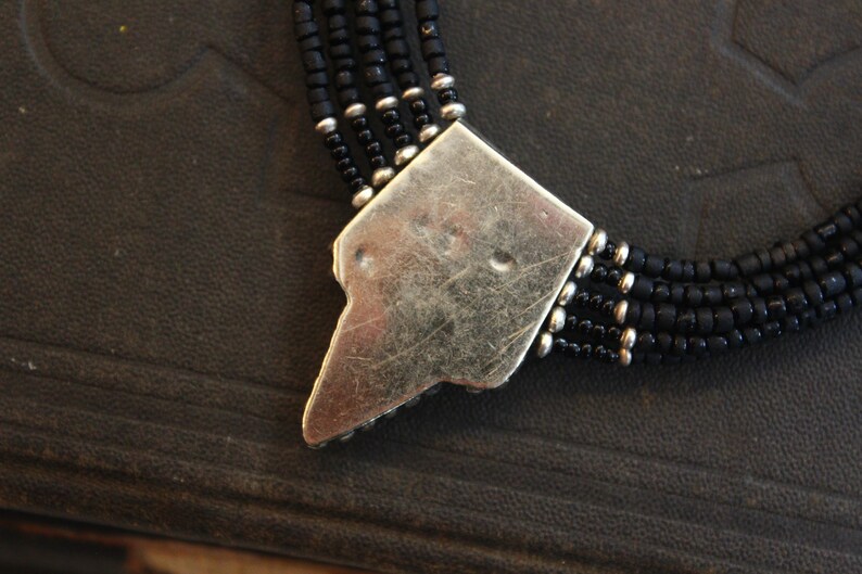 May include: Close-up of a black beaded necklace with a silver-colored clasp. The necklace features multiple strands of small, dark beads. The clasp is a geometric shape with a worn, textured appearance, and is connected to the beaded strands.