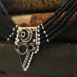 May include: A multi-strand black beaded necklace with a silver-tone pendant. The pendant features black and white circular and triangular designs. The necklace is displayed on a stack of antique books.