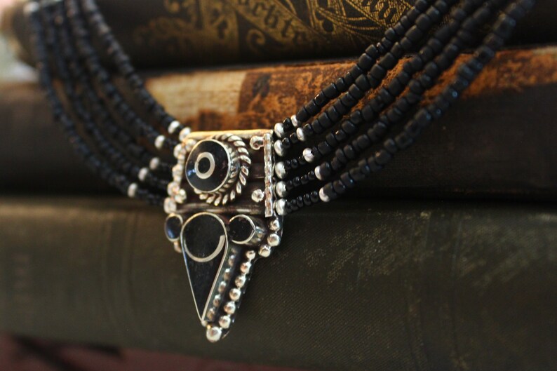 May include: A multi-strand black beaded necklace with a silver-tone pendant. The pendant features black and white circular and triangular designs. The necklace is displayed on a stack of antique books.