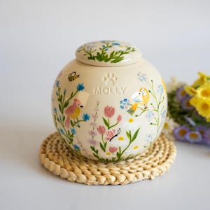 May include: A round, cream-colored ceramic urn with a matching lid, featuring hand-painted illustrations of colorful flowers, birds, a bee, and a butterfly. The name MOLLY is embossed in the center above a small paw print icon.