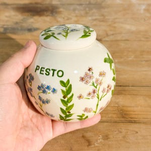 May include: A white ceramic pesto jar with a lid, decorated with blue and pink floral patterns and green leaves. The word "PESTO" is written in green on the jar's front. The jar is held in a hand.