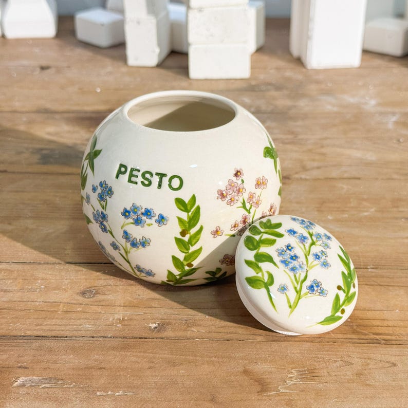 May include: A white ceramic pesto jar with a matching lid. The jar is round with the word "PESTO" in green lettering. It is decorated with blue and pink floral designs and green leaves. The lid is oval and similarly decorated.