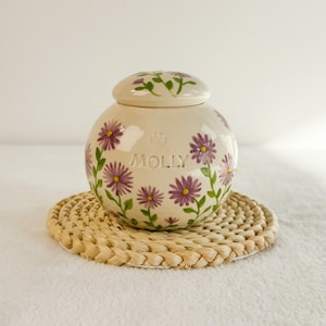 May include: A cream-colored ceramic pet urn with a lid, adorned with purple daisy floral designs and the name "MOLLY". The urn sits on a woven coaster. The design includes a paw print.