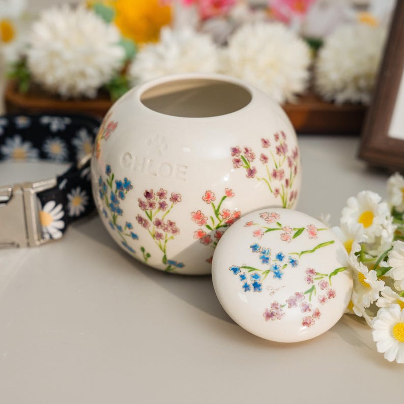 May include: A white ceramic pet urn with a lid, adorned with delicate floral patterns in pink, blue, and green. The urn is personalized with the name "CHLOE". A daisy-patterned pet collar is visible in the background.