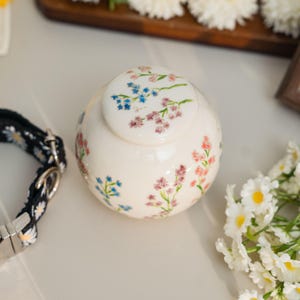 May include: A cream-colored ceramic urn with a lid, adorned with delicate floral patterns in shades of blue, pink, and red. The urn has a rounded shape and is decorated with painted flowers. A black and white daisy collar is visible in the image.