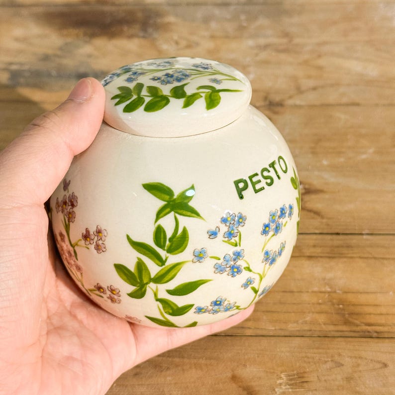May include: A round, off-white ceramic jar with a lid, decorated with blue forget-me-nots and green leaves. The word "PESTO" is written in green. The jar is held in a hand.