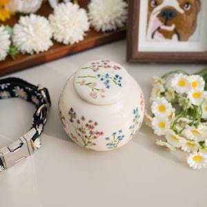 May include: A cream-colored pet urn with a floral design in pink, blue, and purple. The urn has a matching lid and is next to a black and white daisy-patterned dog collar and a bouquet of white flowers.