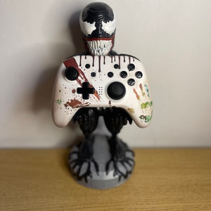 May include: A white video game controller with brown and red splattered paint, held by a black Venom figurine. The controller has a white plus sign and a black circle button.