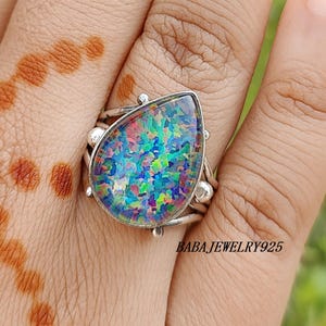 May include: A silver ring with a large, teardrop-shaped opal stone. The opal displays a vibrant array of colors, including blue, green, and purple. The ring has a detailed silver band with decorative elements. The text "BABAJWELRY925" is visible.