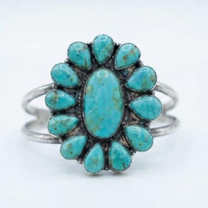 May include: A silver cuff bracelet featuring a large central oval turquoise stone surrounded by twelve smaller teardrop-shaped turquoise stones. The metal band is split into two parallel wires that meet at the sides of the stone cluster setting.