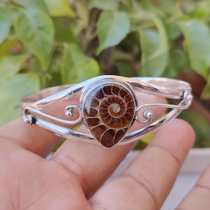 Pode incluir: Um bracelete de prata com uma pedra preciosa de amonite marrom. A amonite é um fóssil de concha em espiral com um centro marrom claro e bordas externas marrom escuro.