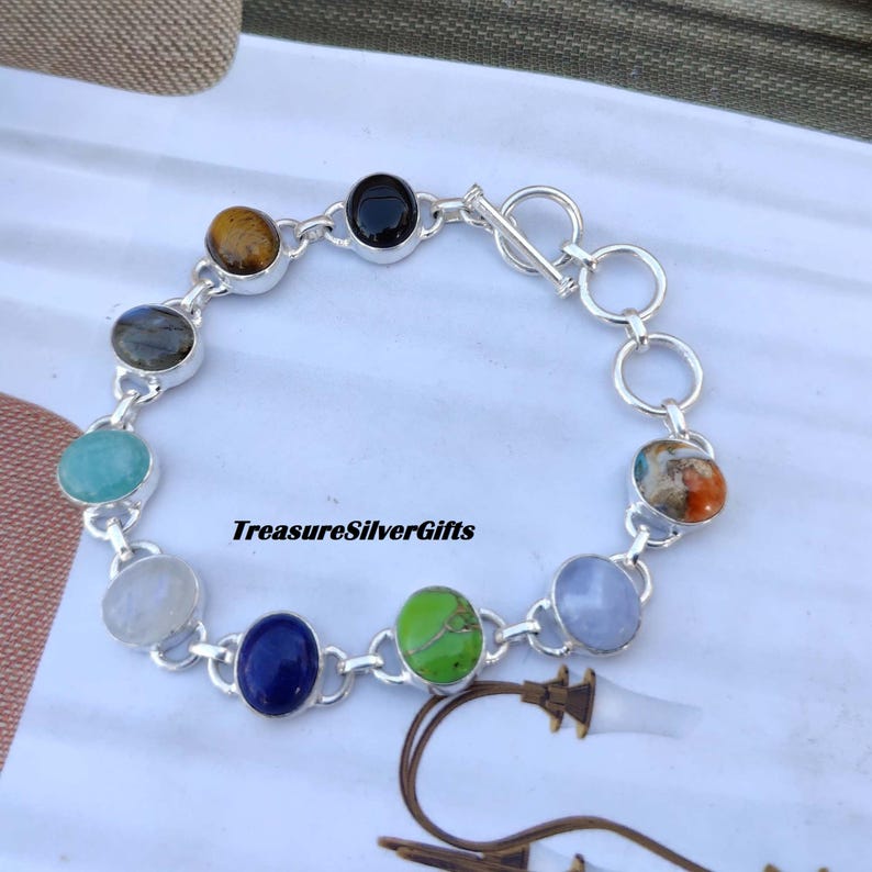 May include: A silver bracelet with a chain link design and nine oval-shaped gemstones. The gemstones are various colors, including black, brown, green, blue, and white.