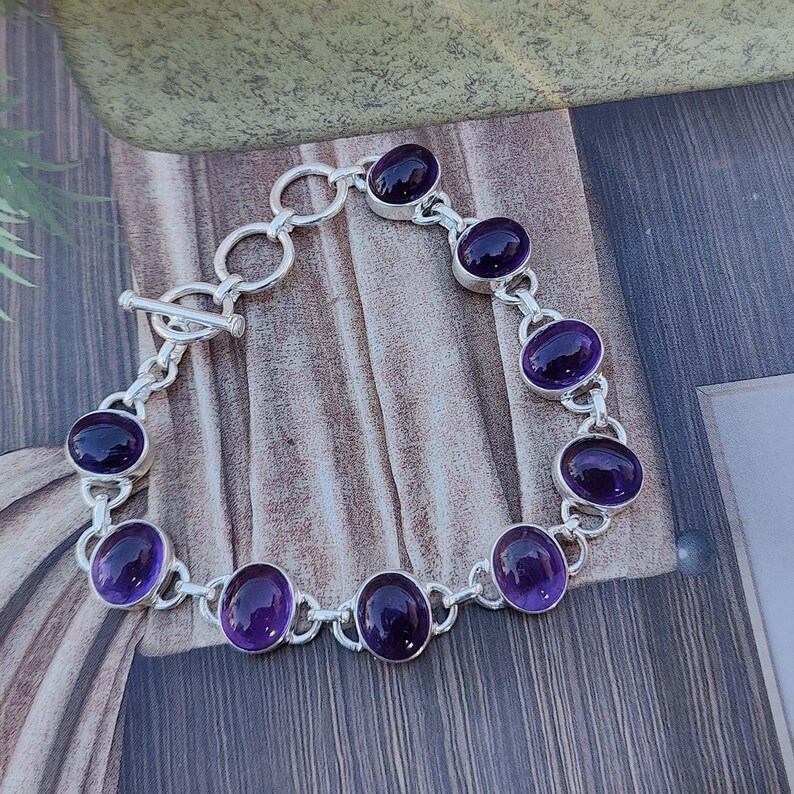 K&ouml;nnte beinhalten: Ein silbernes Kettenarmband mit sieben ovalen Amethyst-Edelsteinen. Das Armband hat einen Schiebeverschluss.