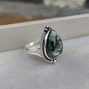 Puede incluir: Un anillo de plata con una piedra preciosa verde y blanca en forma de pera. El anillo tiene una banda de plata detallada con un diseño de cuerda.