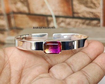 May include: A silver bangle bracelet with a pink and orange gemstone set in the center. The bracelet is open and has a simple design.