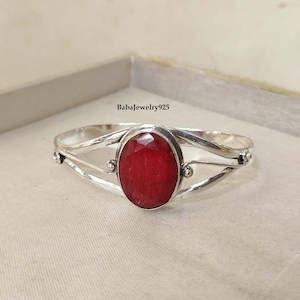 May include: A silver bangle bracelet with a red oval gemstone. The bracelet has a delicate, twisted design and is accented with small silver beads.