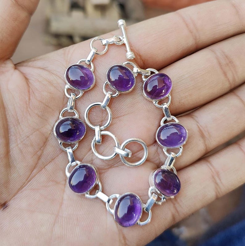 K&ouml;nnte beinhalten: Ein silbernes Armband mit einem Schiebeverschluss verf&uuml;gt &uuml;ber acht ovale Amethyst-Edelsteine. Das Armband befindet sich auf einer Hand.