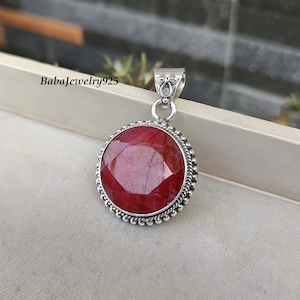 Puede incluir: Un colgante de plata con una gran piedra de rubí roja redonda. El colgante tiene un borde de plata detallado y una anilla para sujetarlo a una cadena.