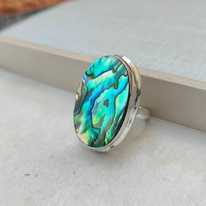 Puede incluir: Un anillo de plata con una gran incrustación ovalada de concha de abulón. La concha tiene un patrón azul, verde e iridiscente.