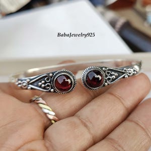 May include: Silver bangle bracelet with two red gemstone accents. The bracelet features a delicate floral design.