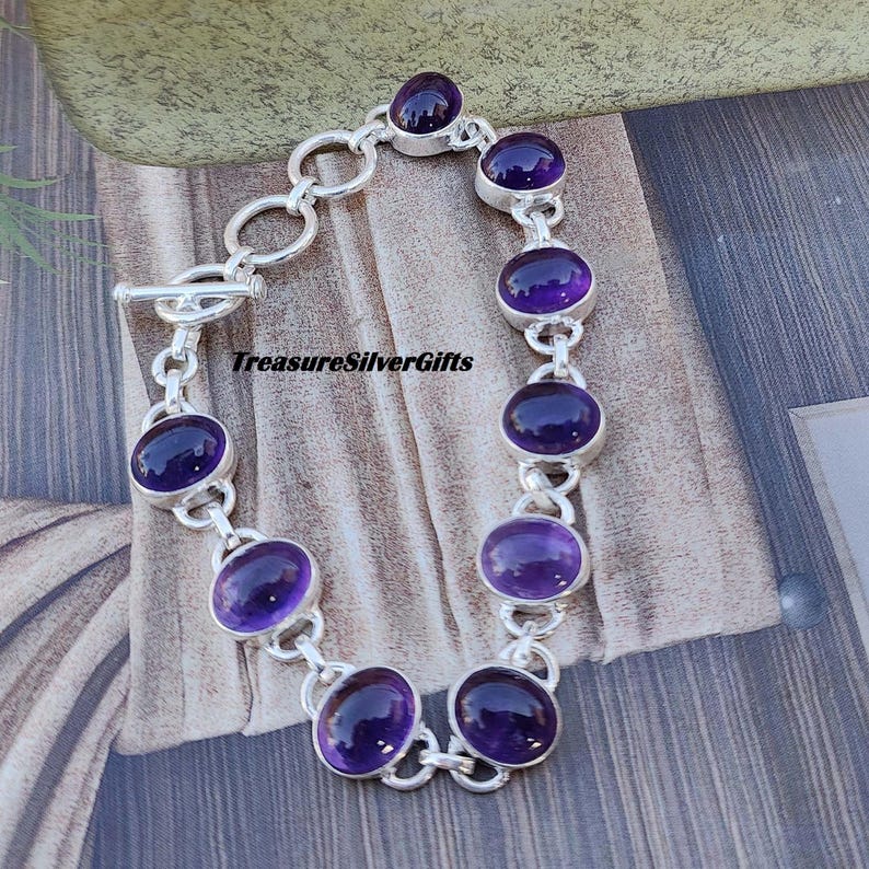 K&ouml;nnte beinhalten: Ein silbernes Armband mit einem Schiebeverschluss und acht violetten Amethyst-Edelsteinen. Das Armband liegt auf einer Holzoberfl&auml;che.