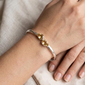 May include: A silver cuff bracelet with two yellow, teardrop-shaped gemstones. The bracelet is worn on a wrist, highlighting the polished metal and the warm hues of the stones. The design is simple and elegant.