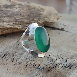 May include: A silver ring with a green oval gemstone set in a unique, open design. The ring is on a wooden surface.