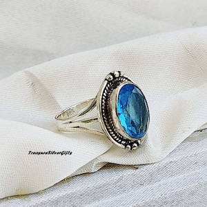 Pode incluir: Um anel de prata com uma grande pedra preciosa oval azul brilhante. A pedra preciosa é definida em uma moldura detalhada com uma borda semelhante a uma corda. O texto "TreasureSilverGifts" é visível.