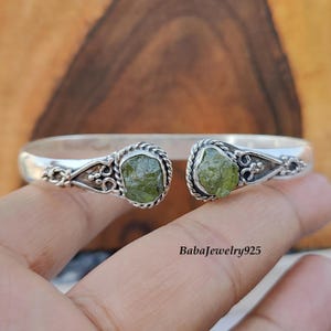 May include: A silver cuff bracelet with two raw, light green peridot gemstones. The peridot stones are set in a decorative, open-ended design with intricate detailing. The bracelet is shown on a hand.