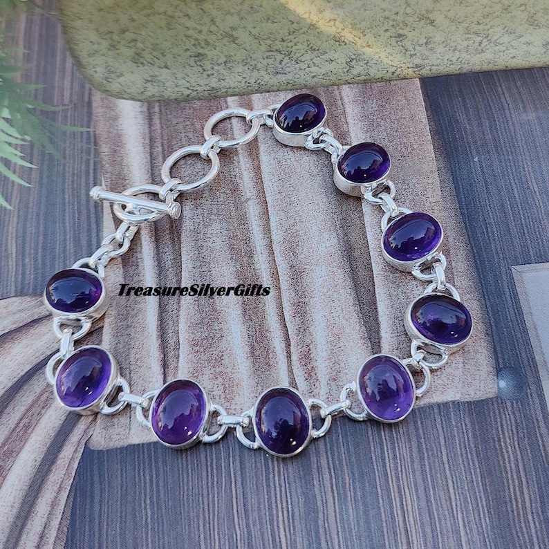 K&ouml;nnte beinhalten: Ein silbernes Armband mit einem Gliederketten-Design und ovalen Amethyst-Edelsteinen. Das Armband hat einen Schiebeverschluss.
