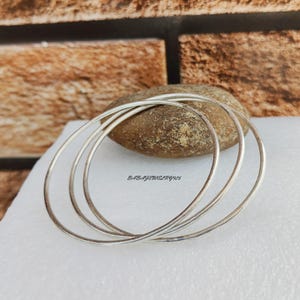 Puede incluir: Tres pulseras de plata apiladas una encima de la otra. Las pulseras son lisas y brillantes.