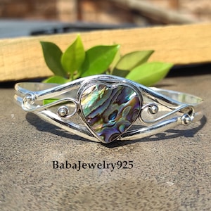 Puede incluir: Una pulsera plateada con un centro en forma de corazón de concha de abulón. La concha iridiscente muestra una mezcla de tonos verdes, azules y morados. La pulsera presenta remolinos decorativos y el texto "BabaJewelry925".