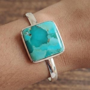 May include: A silver cuff bracelet featuring a large, square-shaped turquoise stone with natural veining in shades of bright blue and teal. The polished metal band has a smooth, solid finish and connects to the stone setting on both sides.
