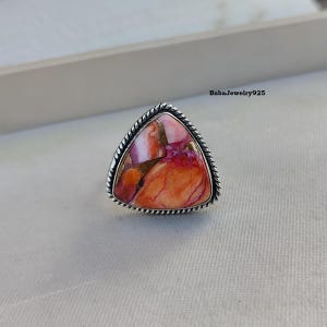May include: A silver ring with a triangular orange, pink, and white gemstone. The ring has a rope-like design around the edge of the setting.