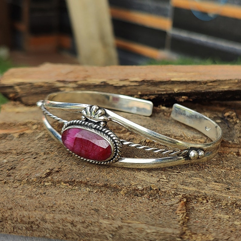 May include: A silver bangle bracelet with a pink oval gemstone set in a silver bezel. The bracelet has a twisted wire design and a decorative silver accent.