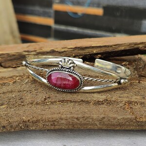 May include: A silver bangle bracelet with a red gemstone set in a silver bezel. The bracelet has a twisted wire design and a decorative floral accent.