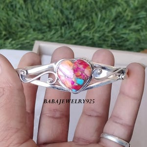 May include: A silver bangle bracelet with a heart-shaped pink, orange, and turquoise gemstone. The bracelet has a delicate, swirling design.