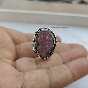 Op de afbeelding: Een zilveren ring met een roze en groene edelsteen gezet in een zilveren bezel. De ring wordt gedragen aan een vinger.