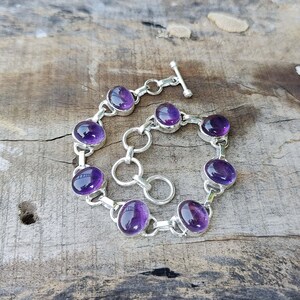 K&ouml;nnte beinhalten: Ein silbernes Kettenarmband mit ovalen Amethyst-Edelsteinen. Das Armband hat einen Karabinerverschluss.
