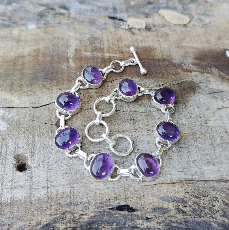 K&ouml;nnte beinhalten: Ein silbernes Kettenarmband mit ovalen Amethyst-Edelsteinen. Das Armband hat einen Karabinerverschluss.